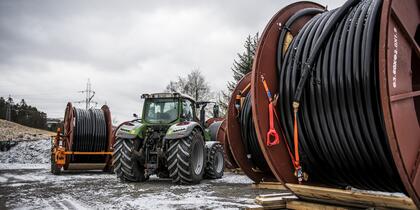 Store tromler med svart kabel er satt i ring rundt en grønn traktor som blir liten mellom tromlene. 